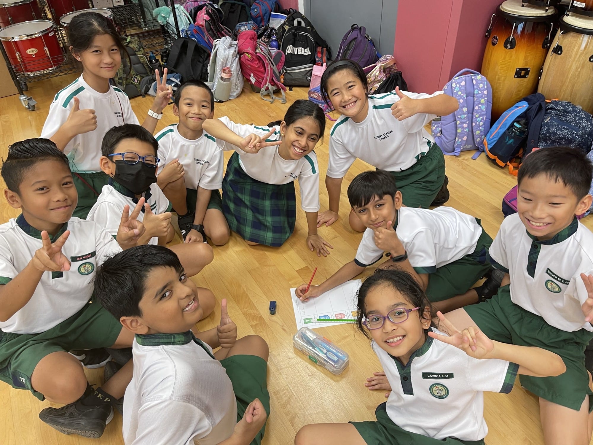 Group of smiling school children in white and green uniforms sitting on a floor, posing with peace signs and thumbs up.